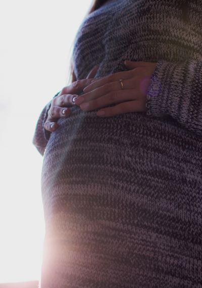 Schwangere Frau im Pullover, die ihren Bauch berührt. Im Hintergrund scheint die Sonne.